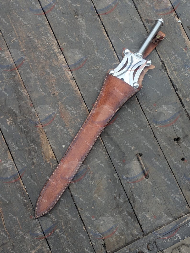 May include: A medieval-style sword with a silver blade and handle, in a brown leather sheath. The sword's hilt has a decorative design. The sheath is stitched with white thread. The sword rests on a weathered wooden surface.