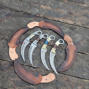 May include: A collection of curved karambit knives with ornate handles and Damascus steel blades, displayed with brown leather sheaths. The knives are arranged on a weathered wooden surface, showcasing their craftsmanship.