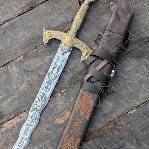 Könnte beinhalten: Ein verziertes Schwert mit silberner Klinge und goldenen Akzenten, mit einem roten Edelstein auf dem Knauf. Die Klinge ist mit dem Namen "Killian Jones" graviert. Eine braune Lederscheide mit dekorativem Text und Riemen ist dabei.