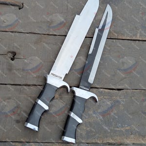Könnte beinhalten: Zwei silber-schwarze Messer mit dem Wort "PREDATOR" auf der Klinge. Die Messer haben schwarze Griffe mit silbernen Akzenten. Die Klingen sind lang und scharf, und die Messer werden auf einer Holzoberfläche präsentiert.