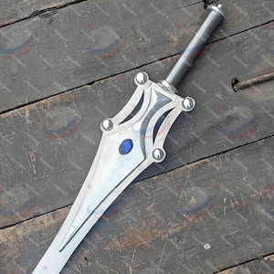 May include: A silver sword with a decorative hilt featuring a blue gem and rounded accents. The blade is long and pointed, with a central ridge. The sword rests on a weathered wooden surface.