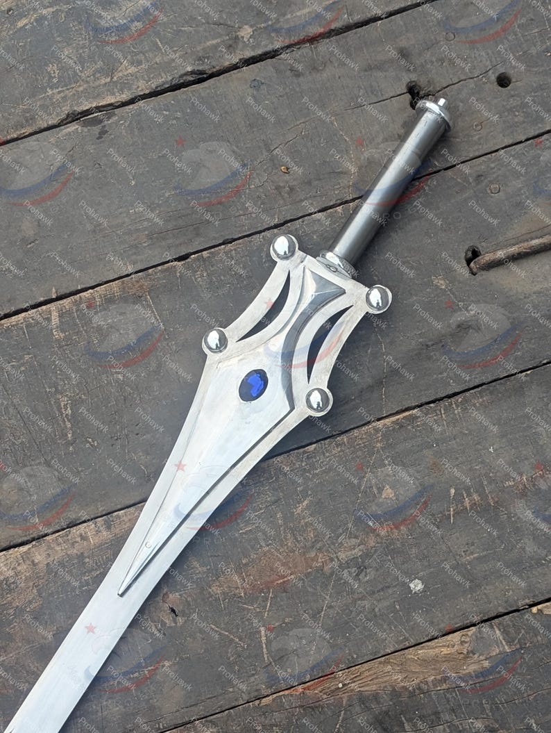 May include: A silver sword with a decorative hilt featuring a blue gem and rounded accents. The blade is long and pointed, with a central ridge. The sword rests on a weathered wooden surface.