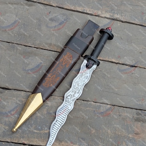 May include: A decorative dagger with a silver blade and black handle, resting beside its brown leather sheath. The sheath has gold accents and a decorative design. The dagger's handle is topped with a red gem.