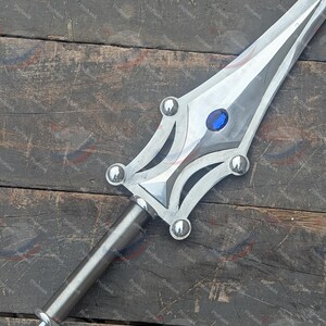 May include: A silver sword with a polished blade and a decorative hilt. The hilt features a blue gem and rounded silver accents. The sword is lying on a wooden surface.