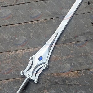 May include: A silver sword with a blue gem on the hilt. The blade is long and pointed, and the handle features decorative details. The sword is displayed on a weathered wooden surface.