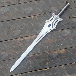 May include: A silver sword with a long, pointed blade and a decorative hilt. The hilt features a blue gem and silver accents. The sword is displayed on a weathered wooden surface.