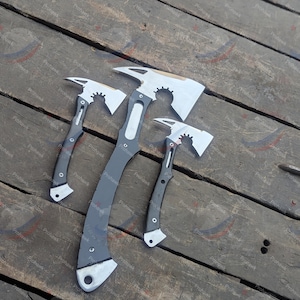 May include: Three axes with silver blades and dark gray handles are arranged on a weathered wooden surface. The axes vary in size, with a curved design and a hole at the base of the handle. The blades have a serrated edge.