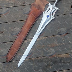 May include: A silver sword with a decorative hilt and a blue gem, next to a brown leather sheath. The sword has a long, pointed blade. The sheath has a strap for carrying. The items are on a wooden surface.