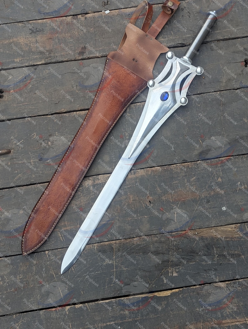 May include: A silver sword with a decorative hilt and a blue gem, next to a brown leather sheath. The sword has a long, pointed blade. The sheath has a strap for carrying. The items are on a wooden surface.