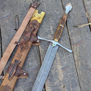 Könnte beinhalten: Ein Damaststahl-Schwert mit gemusterter Klinge, einem silbernen Parierstange mit kleinem grünen Stein und einem Holzgriff mit detaillierten Schnitzereien. Dazu gehört eine passende Holzscheide mit Lederriemen und einer Metallschnalle.