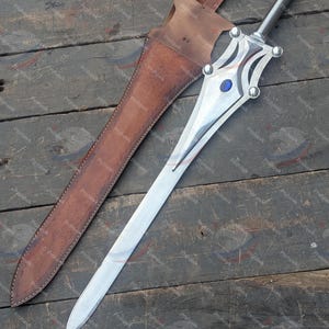 May include: A silver sword with a decorative hilt and a blue gem, next to a brown leather sheath. The sword has a long, pointed blade and a detailed handle with silver accents. The sheath has a strap for carrying.