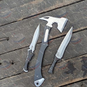 May include: Three metal weapons: a silver and black axe with a toothed wheel design, and two knives. One knife has a curved blade with a black handle and the other has a straight blade with a black handle.