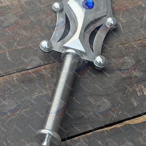 May include: A close-up of a silver sword with a blue gem in the center of the blade. The handle is cylindrical and the blade has decorative cutouts and rounded accents. The sword is set against a wooden surface.