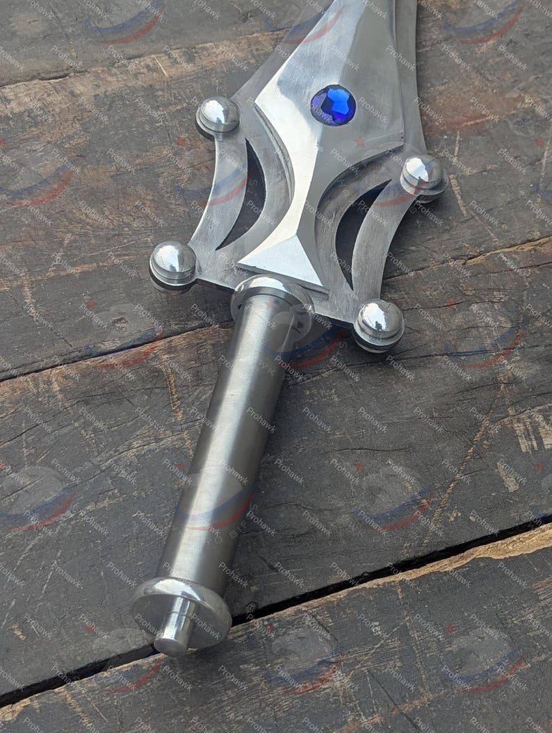 May include: A close-up of a silver sword with a blue gem in the center of the blade. The handle is cylindrical and the blade has decorative cutouts and rounded accents. The sword is set against a wooden surface.