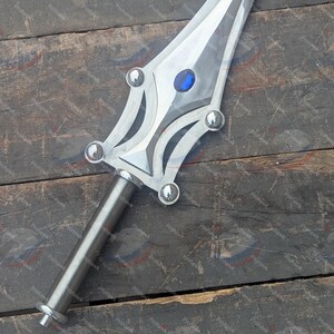 May include: A silver sword with a decorative hilt and blade. The blade has a central groove and a blue gem near the hilt. The hilt features rounded silver accents. The sword is displayed on a weathered wooden surface.