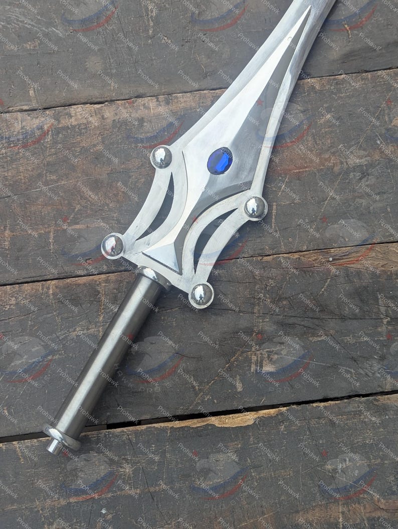 May include: A silver sword with a decorative hilt and blade. The blade has a central groove and a blue gem near the hilt. The hilt features rounded silver accents. The sword is displayed on a weathered wooden surface.