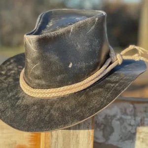 May include: A distressed black leather cowboy hat with a light brown braided band and a knotted tie. The hat has a wide brim and a tall crown, suggesting a rugged, vintage aesthetic.