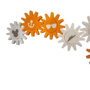 Puede incluir: Colección de adornos decorativos en forma de engranaje en blanco y naranja. Los adornos presentan varios diseños con temática veraniega, incluyendo gafas de sol, un ancla, un barco y una silueta de Mickey Mouse.