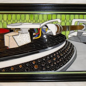 May include: A close-up of a stained glass art piece depicting a turntable. The turntable is made of black, white, and silver glass. The background is a green mosaic of small circles.
