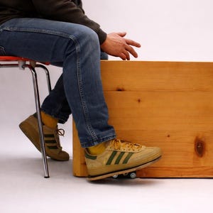 Könnte beinhalten: Eine große, unfertige Holzkiste mit sichtbarer Holzmaserung und Knoten. Die Kiste steht auf kleinen Rädern. Eine Person sitzt auf einem roten Stuhl neben der Kiste und trägt eine blaue Jeans und beige Turnschuhe mit grünen Streifen.