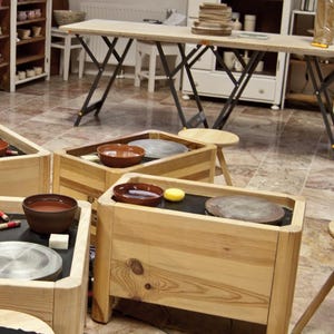 Puede incluir: Un estudio de cerámica con varios tornos de alfarero de madera. Cada torno tiene un cuenco marrón y un disco de metal. El estudio cuenta con una mesa de madera clara y estanterías que exhiben cerámica.