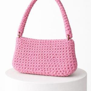 May include: A pink crochet handbag with a braided handle. The bag is rectangular with rounded edges and a textured surface. The handle is attached with gold-colored hardware. The bag is displayed on a white pedestal.