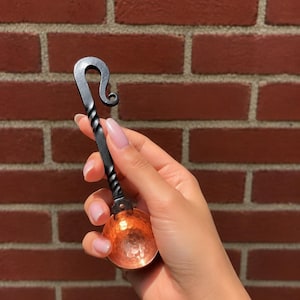 Puede incluir: Una cuchara medidora de metal forjada a mano con un cuenco redondo de cobre martillado y un mango de hierro negro trenzado que termina en un gancho decorativo rizado en la parte superior. La cuchara muestra un contraste entre el cobre y el hierro.