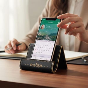 May include: Black leather phone stand with gold stitching and the name "melisa" in gold script. A smartphone displays a calendar app. The stand is on a wooden desk.