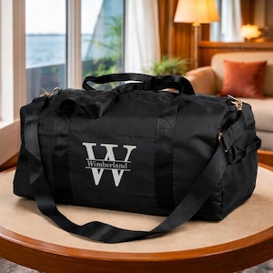 May include: A black duffel bag with a shoulder strap and handles. The bag features the text "Wimberland" in a white, stylized font. The bag is sitting on a round, light-colored table.