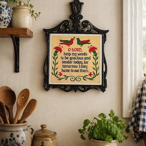 May include: A decorative wall hanging with a black ornate frame. The centre features a cream-coloured tile with a floral and bird design and the text "O LORD, help my words to be gracious and tender today, for tomorrow I may have to eat them."