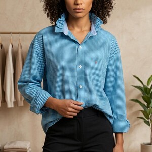 May include: A woman wearing a blue gingham shirt with a light blue collar and cuffs, paired with black pants. The shirt has a small red logo on the chest. The background features a clothing rack and a potted plant.