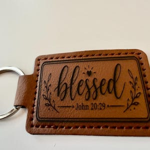 May include: A rectangular brown leather keychain featuring dark brown stitching around the edges. The center is laser-engraved with the word blessed in a cursive script, a small heart icon, and the text John 20:29, flanked by leaf illustrations.