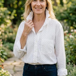 Puede incluir: Una mujer con una camisa blanca con lunares azul marino y vaqueros azul oscuro. La camisa tiene un cuello con volantes y un bolsillo en el pecho. La mujer sonríe en un jardín.