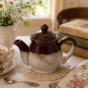 May include: A ceramic teapot featuring a two-tone design with a glossy dark brown upper section and a textured, crackled cream-coloured base. The teapot includes a matching lid with a knob handle, a curved spout, and a sturdy side handle.