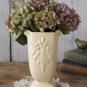 Peut inclure: Un vase en céramique de couleur crème avec un bord évasé et un motif en relief symétrique en forme de feuille sur le devant. Le vase possède une base carrée et est rempli d'un bouquet d'hortensias séchés dans des tons de vert, brun et violet.