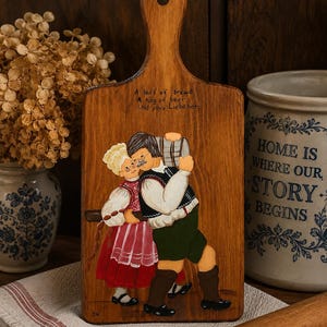 May include: Wooden cutting board with a handle, featuring a hand-painted scene of a couple carrying a beer keg. The board has the text "A loaf of bread, A keg of beer, Und you, Liebchen." A rolling pin and a ceramic jar are in the background.
