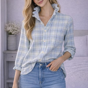 May include: A light blue plaid shirt with a ruffled collar and cuffs, paired with light wash blue jeans. The shirt has a button-down front and a single pocket. The overall style is casual and feminine.