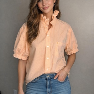 Könnte beinhalten: Pfirsichfarbenes, kurzärmeliges Hemd mit vertikalen weißen Streifen. Das Hemd hat einen gerüschten Kragen und Manschetten, eine Vordertasche und einen Knopfverschluss. Das Hemd wird mit einer blauen Jeans kombiniert.