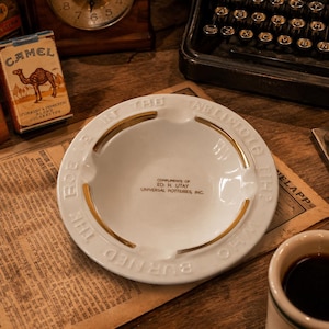 Puede incluir: Cenicero de cerámica blanca con ribete dorado y el texto "BURNED WHO IN THE TABLIREOLE HE". El cenicero también tiene el texto "COMPLIMENTS OF ED. H. UTAY UNIVERSAL POTTERIES, INC.". El cenicero está sobre una superficie de madera.