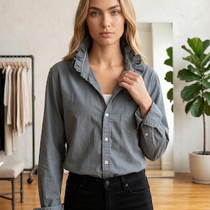 May include: A gray button-down shirt with a ruffled collar and cuffs featuring a white polka dot pattern. The shirt is paired with black jeans. The image is taken indoors with a neutral background.