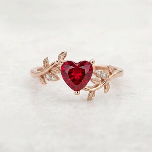 Puede incluir: Un anillo de oro rosa con una piedra preciosa roja central en forma de corazón. La banda está diseñada como una enredadera con hojas texturizadas que se extienden desde el centro, acentuadas por pequeñas piedras claras de corte redondo.