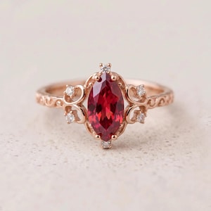 Op de afbeelding: Een roségouden ring met een centrale dieprode edelsteen in marquise-slijpvorm. De zetting is versierd met kleine ronde witte steentjes en ingewikkelde kruldetails langs de ringband, wat zorgt voor een gedetailleerd en sierlijk uiterlijk.