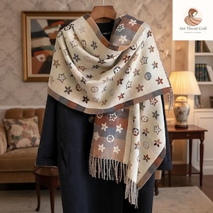 May include: A beige scarf featuring a repeating pattern of brown flowers, stars, and circles, draped over a dark coat. The scarf has a fringed edge and a brown and grey checked border. The scarf is hanging on a wooden hanger.