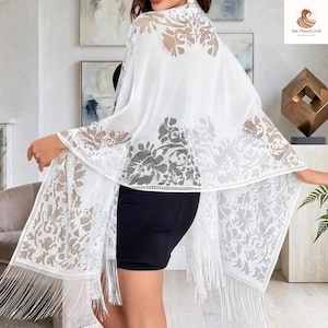 Puede incluir: Un chal de encaje blanco con diseño floral y flecos. El chal se lleva sobre un vestido negro. El delicado diseño calado lo hace adecuado para ocasiones especiales. Fabricado con tejido ligero.