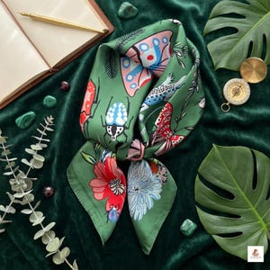 May include: Green silk scarf with a colorful insect and floral pattern. The scarf features a variety of insects, including beetles and butterflies, along with red, pink, and blue flowers. The scarf is knotted and displayed on a green velvet surface.