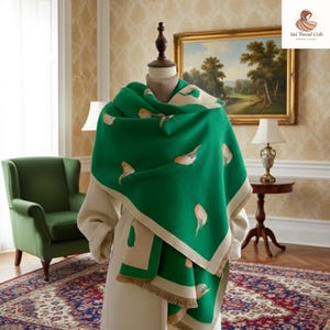 May include: A green scarf with a bird pattern is draped over a mannequin. The scarf has a cream-colored border and fringe. The background includes a painting, a green armchair, and a small table with a lamp.