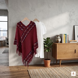 Könnte beinhalten: Ein bordeauxfarbener Poncho mit geometrischem Zierbesatz und Fransen hängt an einem Holzbügel. Daneben hängt ein weißes Kleid. Die Artikel werden vor einer grauen Wand ausgestellt, mit einem Holzschrank und einer Topfpflanze im Hintergrund.
