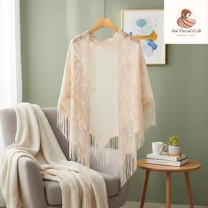 Puede incluir: Un chal de encaje beige con motivos florales y flecos cuelga de una percha de madera. El chal está drapeado sobre un sillón gris con una manta color crema. El logotipo "Aini Thread Craft" es visible.