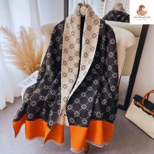 May include: A black and cream-colored scarf with a floral pattern and orange trim. The scarf is draped over a chair and has a fringed edge. The scarf is made of a soft material and is suitable for cool weather.