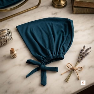 May include: A deep teal fabric bonnet featuring a gathered elastic back and two long fabric ties at the base. The bonnet is constructed from a solid-coloured, smooth material and is designed to be tied securely under the chin or at the nape.